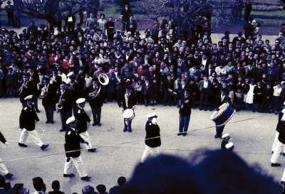 Plano entero de ejército de la Fuerza Naval desfilando junto a una banda tocando instrumentos. A los costados gran cantidad de personas observando en calle aledaña a plaza de la Concordia de La Unión