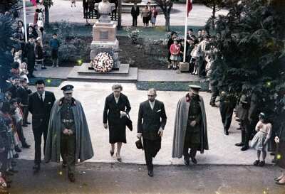 Plano entero de Gobernadora Olga Boettcher junto a 2 militares y dos hombres con Traje caminando frente a estatua de Bernardo O'higgins en plaza de la Concordia de La Unión