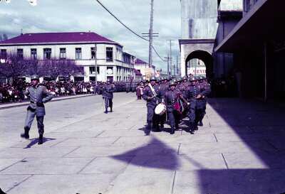 Plano entero de banda de guerra desfilando por calzada de calle Prat frente a locales comerciales de La Unión