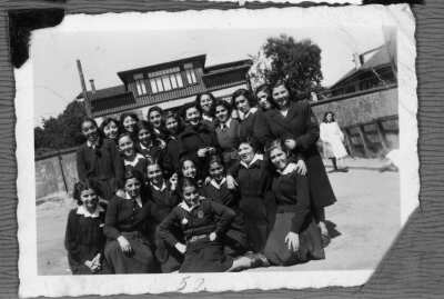 Retrato grupal de niñas adolescentes que se encuentran uniformadas. Están en el interior de un recinto y al fondo se aprecia una casa.