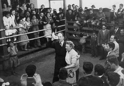 plano general de Olga Boettcher y un hombre vestido de Traje, realizando el paseo durante un baile de cueca. A su alrededor, un grupo numeroso de hombres, mujeres, Niñas y Niños aplauden como es costumbre para avivar el baile. Se encuentran en el interior de un recinto no identificado