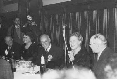 Plano entero de Olga Boettcher sentada junto a tres hombres y una mujer detrás de una mesa. Todos están sonriendo mientras un hombre parado en el fondo está tomando una fotografía. Están en una infraestructura no identificada durante un banquete