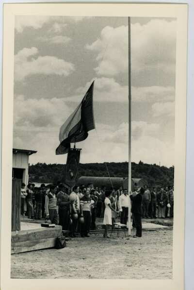 plano general de un hombre y una mujer izando una bandera chilena en el mástil. En el fondo, un grupo de Hombres y Niños se encuentran parados durante una actividad realizada en el interior de la Compañía Carbonífera San Pedro de Catamutún
