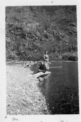 Retrato de plano entero de dos mujeres posando. Una está sentada y la otra parada sobre un mismo tronco, el cual está ubicado a orillas de una playa no identificada