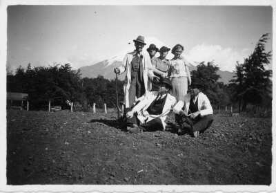 Retrato grupal de plano entero de mujeres y hombres posando. El primer hombre parado de izquierda a derecha tiene consigo una escopeta. En el fondo hay un volcán no identificado.