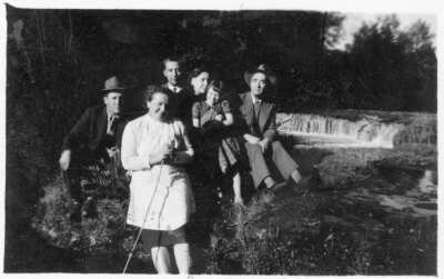Retrato grupal de plano entero de Hombres, Mujeres y una niña posando sentados en el pasto. En el fondo hay una pequeña cascada no identificada.