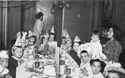 Retrato de plano entero de Niños y Niñas posando alrededor de una mesa durante la celebración de una fiesta de cumpleaños. Les acompañan dos mujeres quienes están paradas cercas de ellos. Se encuentran en el interior de una casa
