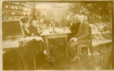 Retrato grupal de plano entero de hombres y dos Carabineros posando sentados alrededor de una mesa. En el fondo se puede observar una bandera chilena y al costado de la mesa un toca disco. Se encuentran en el patio de una casa