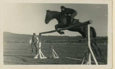 Plano entero de Olga Boettcher realizando una posición de salto ecuestre sobre una valla. A un costado un hombre con sombrero la observa. Lugar no identificado.