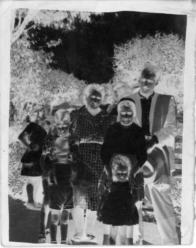 Retrato de plano entero en negativo de una mujer, un hombre, dos niñas y dos niños posando de pie. A sus espaldas hay árboles y arbustos.