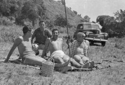 Grupo de Hombres en Traje de Baño posando alrededor de un Asado en la Playa