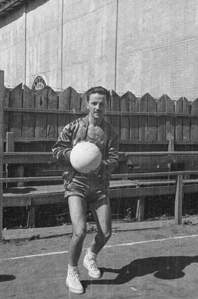 Retrato de plano entero de un jugador de baloncesto, vestido con short y chaqueta, posando con un balón en sus manos.