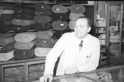 Retrato de plano medio de un hombre posando detrás del mostrador usando un delantal, en el fondo hay un estante de rollos de telas apiladas en una tienda