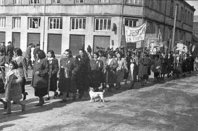 plano general de un grupo de mujeres desfilando, en el medio llevan un lienzo que dice Seccional La escoba La Unión', en el fondo algunas mujeres llevan levantadas escobas y letreros que dicen Ibáñez', un perro va acompañando la marcha que va transitando por una calle durante la campaña presidencial de Carlos Ibáñez del Campo en La Unión