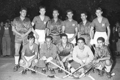 Retrato grupal de plano entero de jugadores de dos equipos de hockey masculino no identificados. En sus poleras algunos tienen grabada la letra N y otros las iniciales CBD. detrás de ellos hay un grupo de Hombres y Mujer que los observa bajo los árboles. El equipo está formado sobre la calle durante la noche