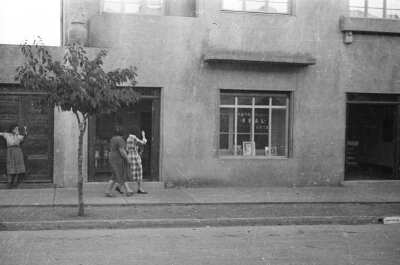 plano general de tres mujeres sobre la vereda. Una se encuentra apoyada en el marco de la puerta de una casa mientras que las otras dos saludan hacia el interior de un estudio fotográfico