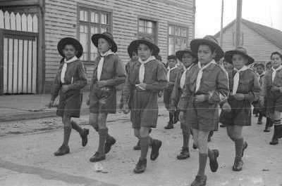 Plano entero de un grupo de Niños pertenecientes a una brigada de scout desfilando en hileras por una calle pavimentada