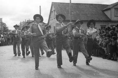 Plano entero de un hombre perteneciente a la brigada de scout, desfilando mientras realiza un saludo hacia su derecha. En el fondo, se encuentra la orquesta sinfónica de la brigada tocando sus instrumentos mientras un grupo numeroso de Niñas, Niños, Mujer y Hombres los observan desfilar por una de las calles de La Unión