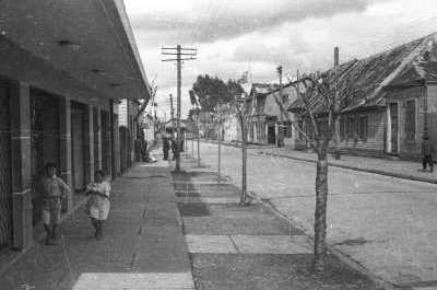 plano general de un niño y un niña caminando descalzos por una vereda en las afueras de una infraestructura. En el fondo, hay un grupo de hombres parados en as afueras de una casa.