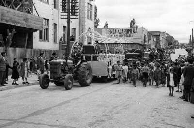 plano general de un hombre conduciendo un tractor, el cual va tirando de una carrocería donde van sentadas un grupo de Niñas junto a la reina de la Escuela superior de niñas Nº2. Le siguen una caravana de autos y también un grupo de Niñas y Niños caminando, en su mayoría, descalzos por la calle pavimentada en el frontis del edificio municipal de La Unión
