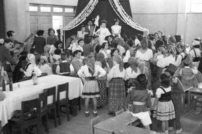 plano general de un grupo de Niñas y Niños disfrazados, parados frente a una tarima, mirando a una Niña vestida de reina. A un costado dos hombres vestidos de Traje tocan sus guitarras, mientras dos mujeres conversan sentadas junto a una mesa. Se encuentran en el gimnasio de la Escuela de Niñas Nº2 durante una actividad escolar