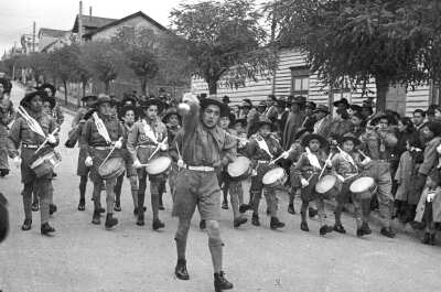 Plano entero del tambor mayor moviendo su guaripola mientras la orquesta sinfónica infantil le sigue tocando sus instrumentos. Todos pertenecen a una brigada de scout no identificada y a un costado, un grupo de Mujer, Niñas y Hombres los observa desfilar por lo largo de una calle pavimentada