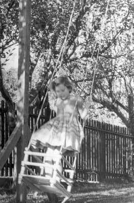 Plano entero de una Niña con Vestido columpiándose bajo un árbo, en el patio de una casa