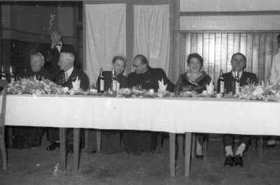 Plano entero de dos mujeres con Vestido una de ellas con corona con 4 Hombres con Trajes sentados alrededor de una mesa.