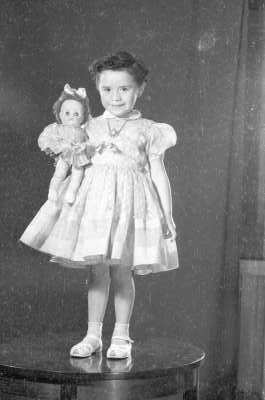 Retrato de estudio de Plano entero de una niña con Vestido posando de frente con su muñeca en brazos. Se encuentra parada sobre una mesa