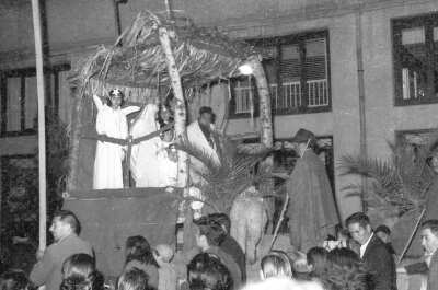 plano general de un grupo de niñas y un hombre reunidos en el interior de un carro alegórico decorado como un pesebre. A los costados Hombres y Mujeres caminando acompañándolo. Se encuentran en las afueras de un edificio de La Unión