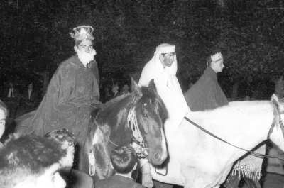 Plano entero de tres hombres disfrazados de reyes magos, paseando sobre sus caballos. Les acompaña un grupo numeroso de personas durante la noche mientras desfilan por las calles de La Unión
