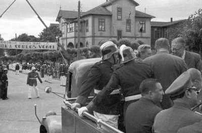Plano entero de Carlos Vogel y un grupo de Hombres y bomberos, en la parte trasera de un camión tres cuartos. En el fondo, un grupo de bomberos se encuentran reunidos bajo un lienzo que dice -bomberos de La Unión dan la bienvenida a Carlos Vogel-. Este homenaje fue realizado durante la llegada del alcalde a la ciudad