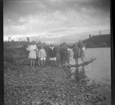 Plano general de ocho personas, niños, niñas y adolescentes, todos posando de pie sobre las piedras en la ribera de un río. Se observa al fondo un puente ferroviario.