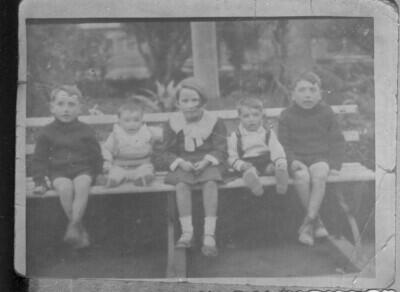 Reproducción de fotografía. Retrato de plano entero de dos niños y una niña, todos de temprana edad junto a dos bebés en el asiento de una plaza no identificada