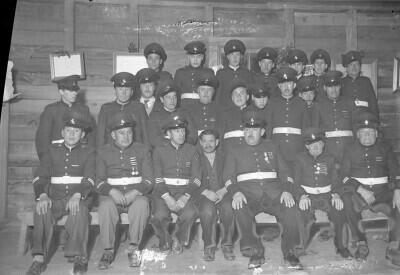 Plano entero de un grupo de hombres vestidos con su uniformes de bomberos acompañados de un hombre mayor con ropa de civil